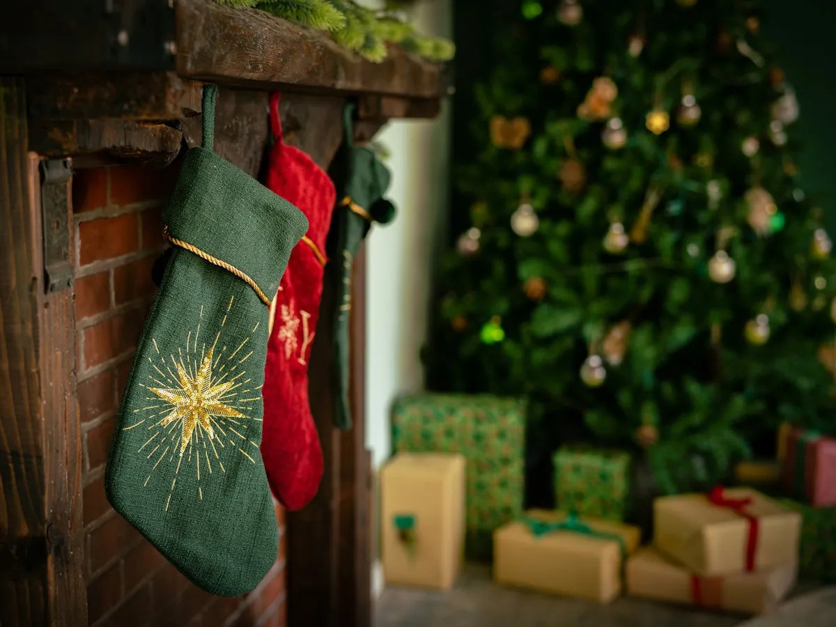 A green embroidered stocking on a brick fireplace with wrapped gifts under the tree