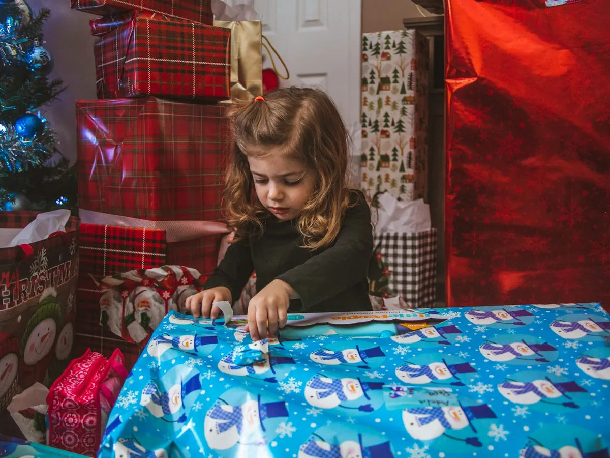 A little girl wrapping a large present in snowman paper among a pile of gifts