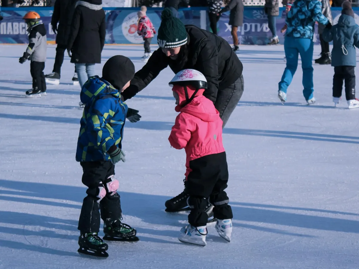 A grown-up helping two small children in helmets learn to ice skate at an outdoor rink