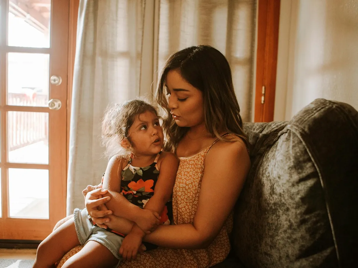 A mother and her little daughter cuddling quietly on a sofa next to a sunny window