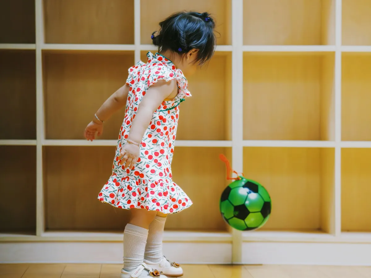 A little girl kicking a green ball indoors in front of wooden shelves