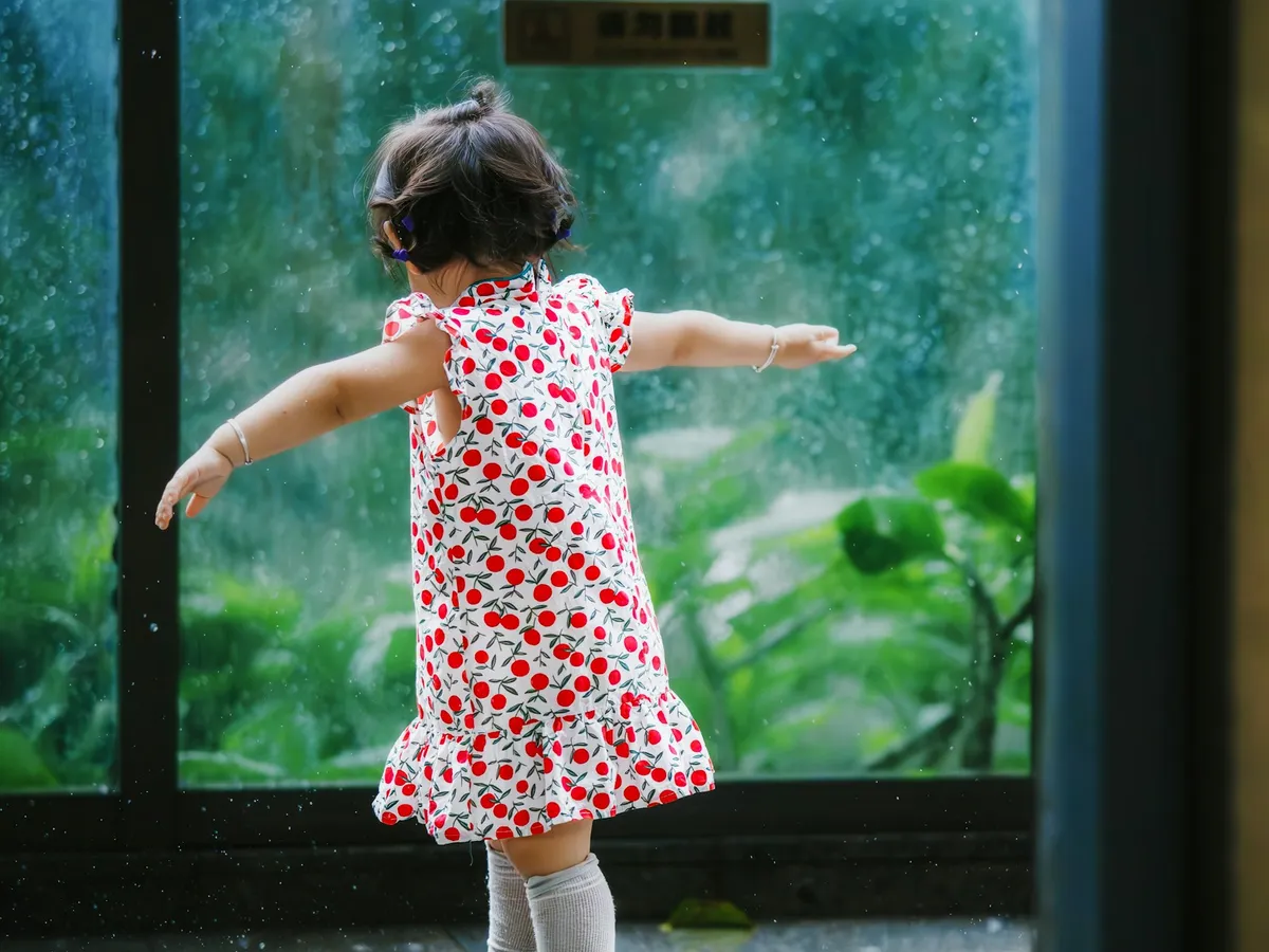 A toddler in a polka-dot dress dancing with arms out by a big window on a rainy day