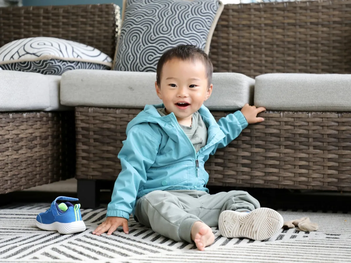 A happy toddler in a blue hoodie sitting on a rug by the sofa, ready to play