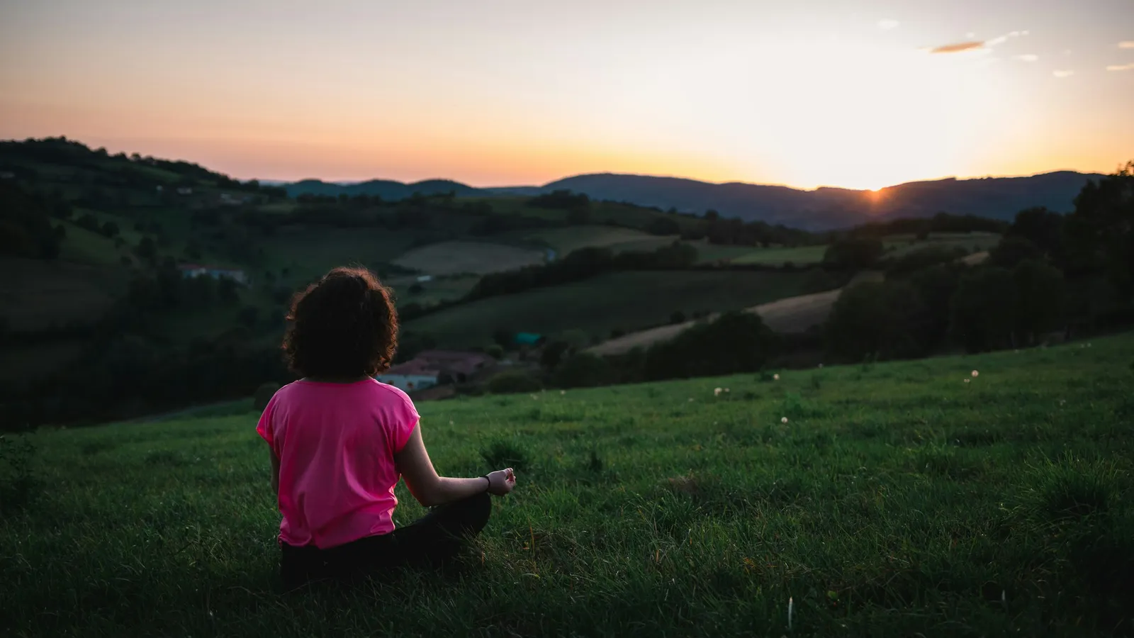 A young girl sitting quietly in the grass, looking out at a soft sunset over the hills