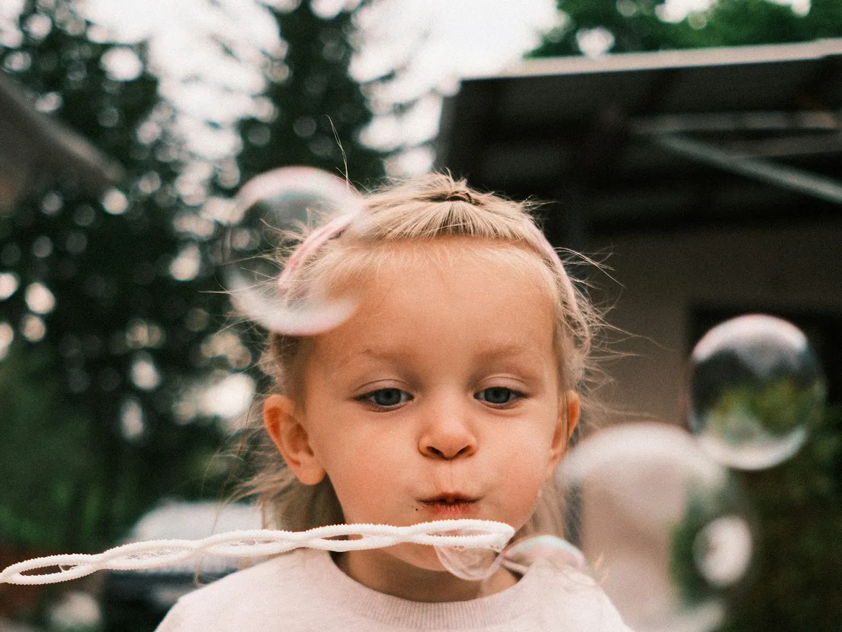 A toddler in a bear sweater blowing a stream of bubbles outdoors