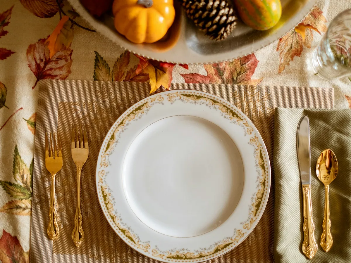 A fall table setting with a gold-rimmed plate on a leaf-print tablecloth, with a small pumpkin and pinecone nearby