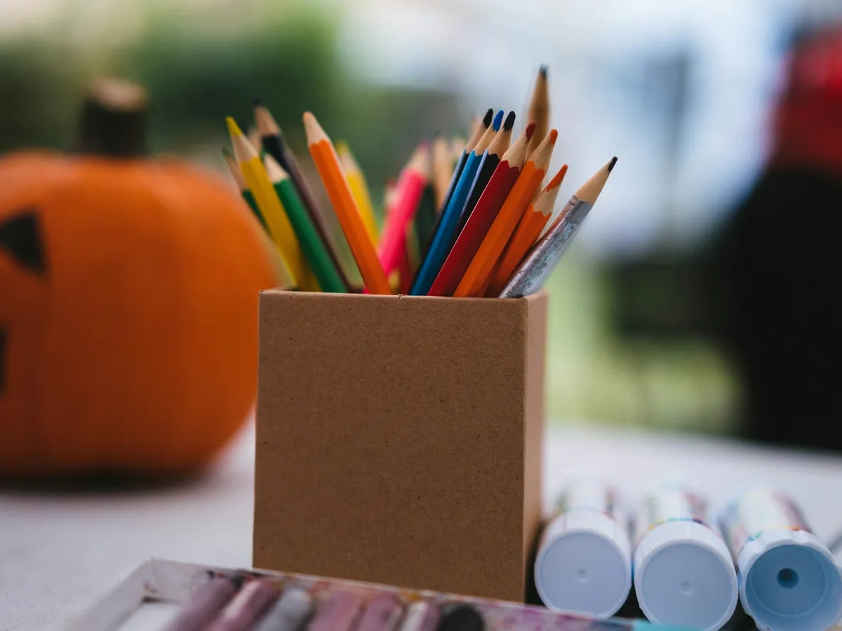A cup of colored pencils and markers on a table beside a carved pumpkin, ready for fall coloring