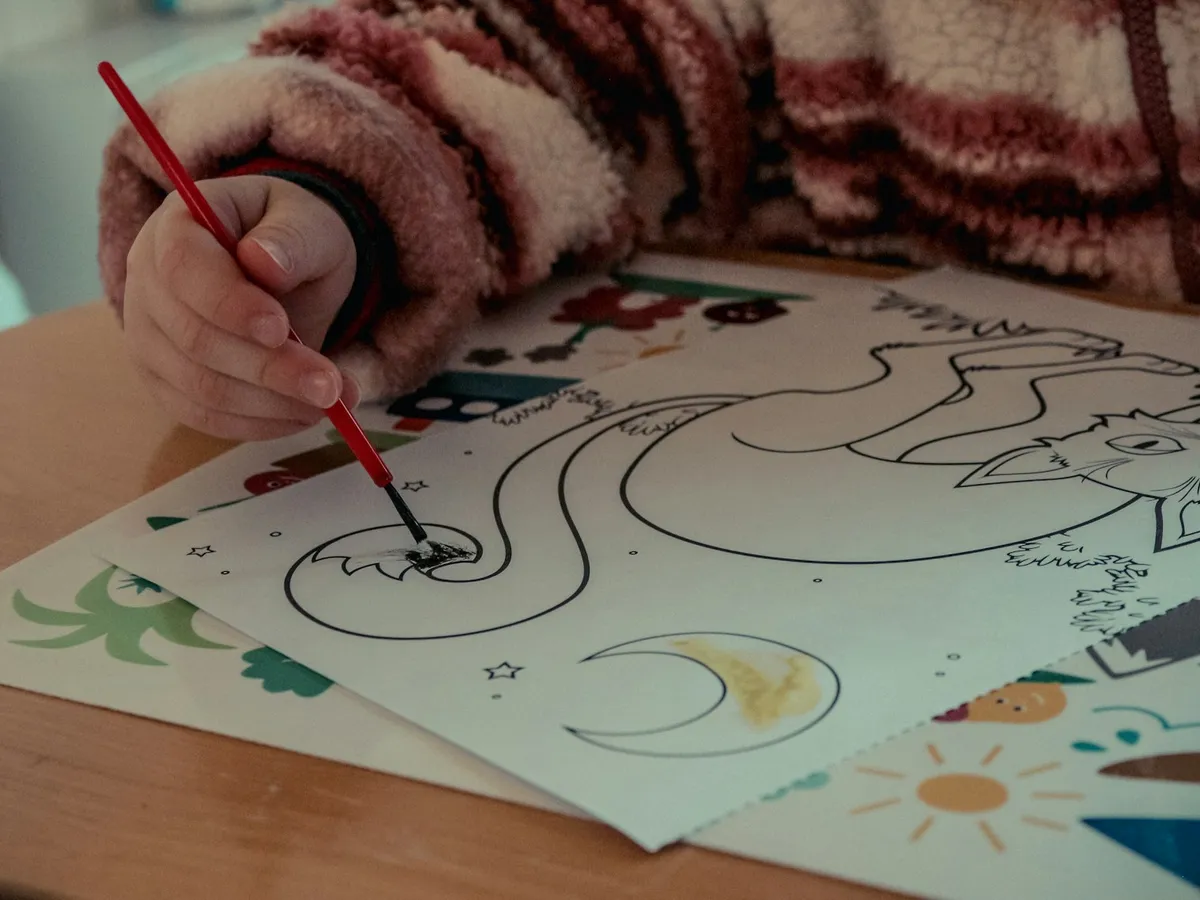 A child’s hand coloring a simple printed coloring page with a red crayon