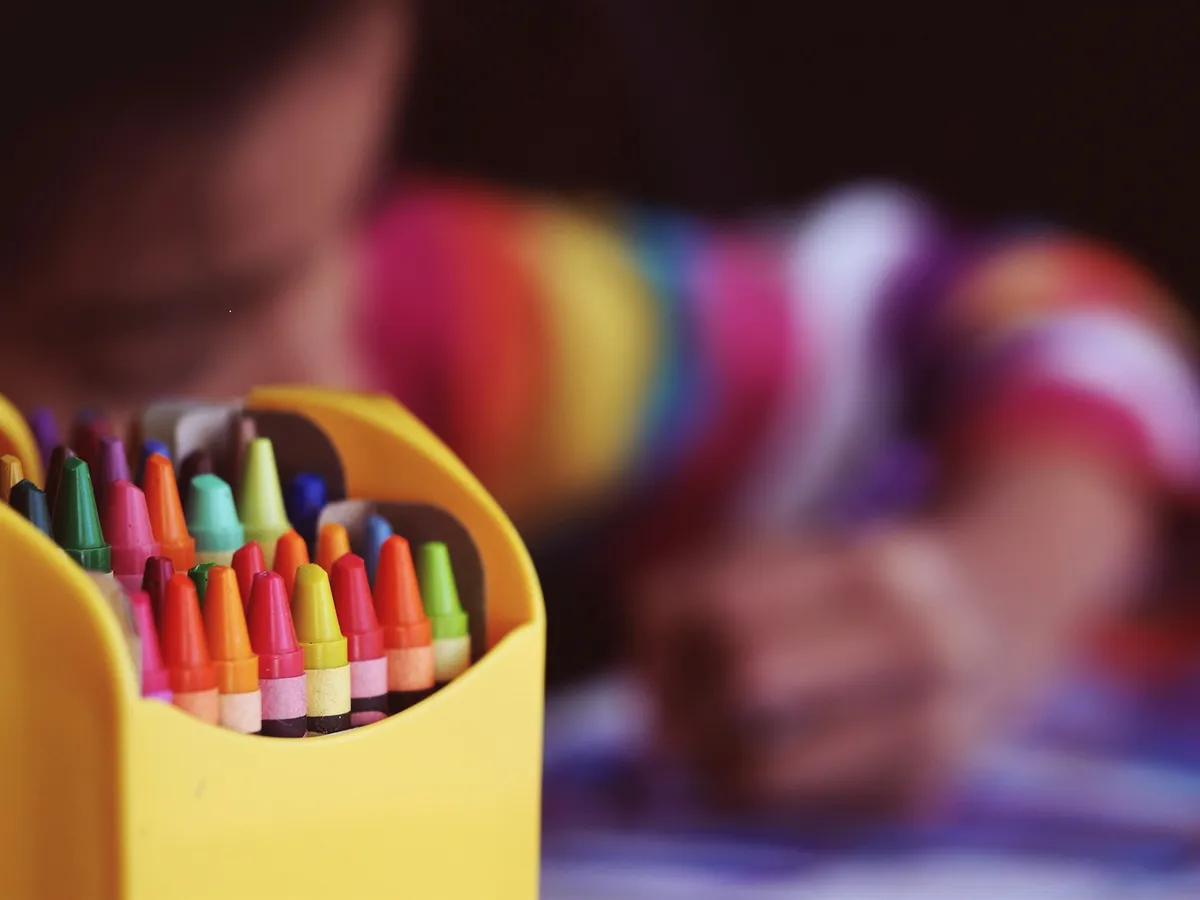 A child coloring behind a yellow box full of bright crayons