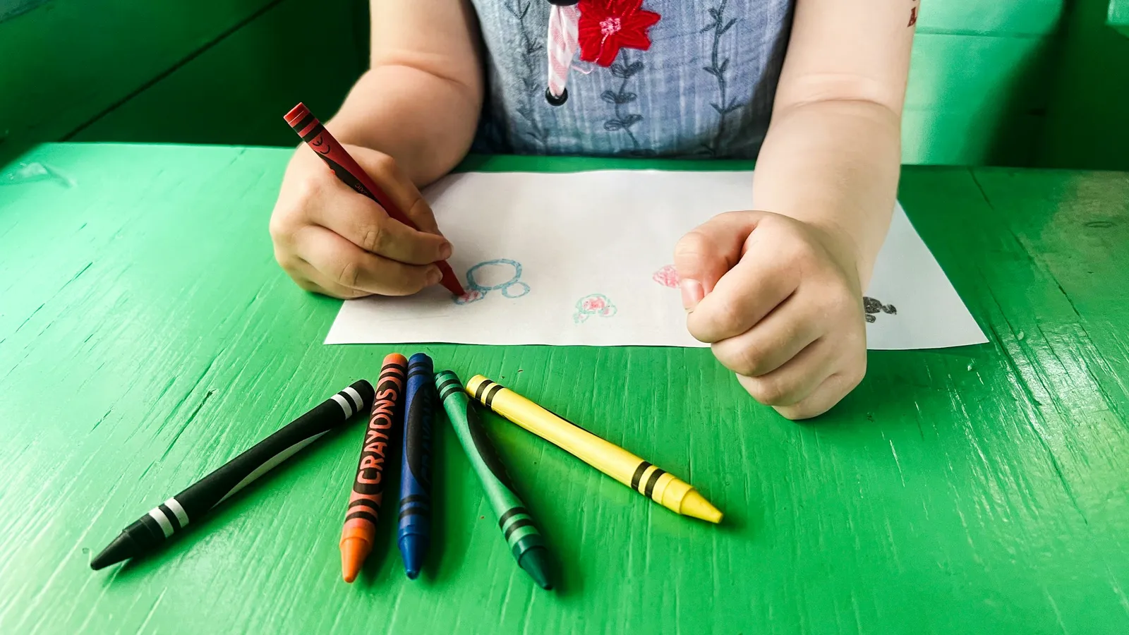 A three-year-old coloring with a red crayon at a bright green table with chunky crayons lined up in front