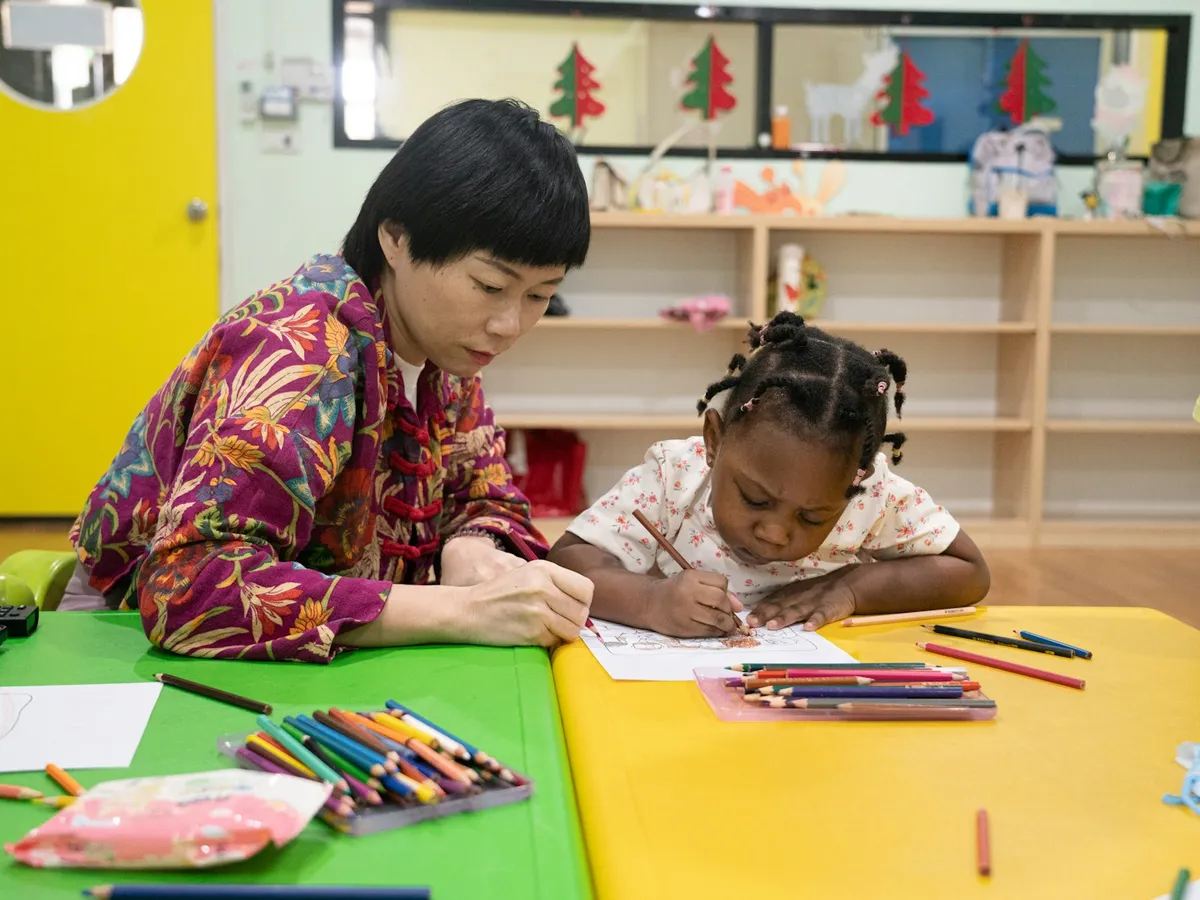 A grown-up and a preschooler coloring side by side at a classroom table with crayons