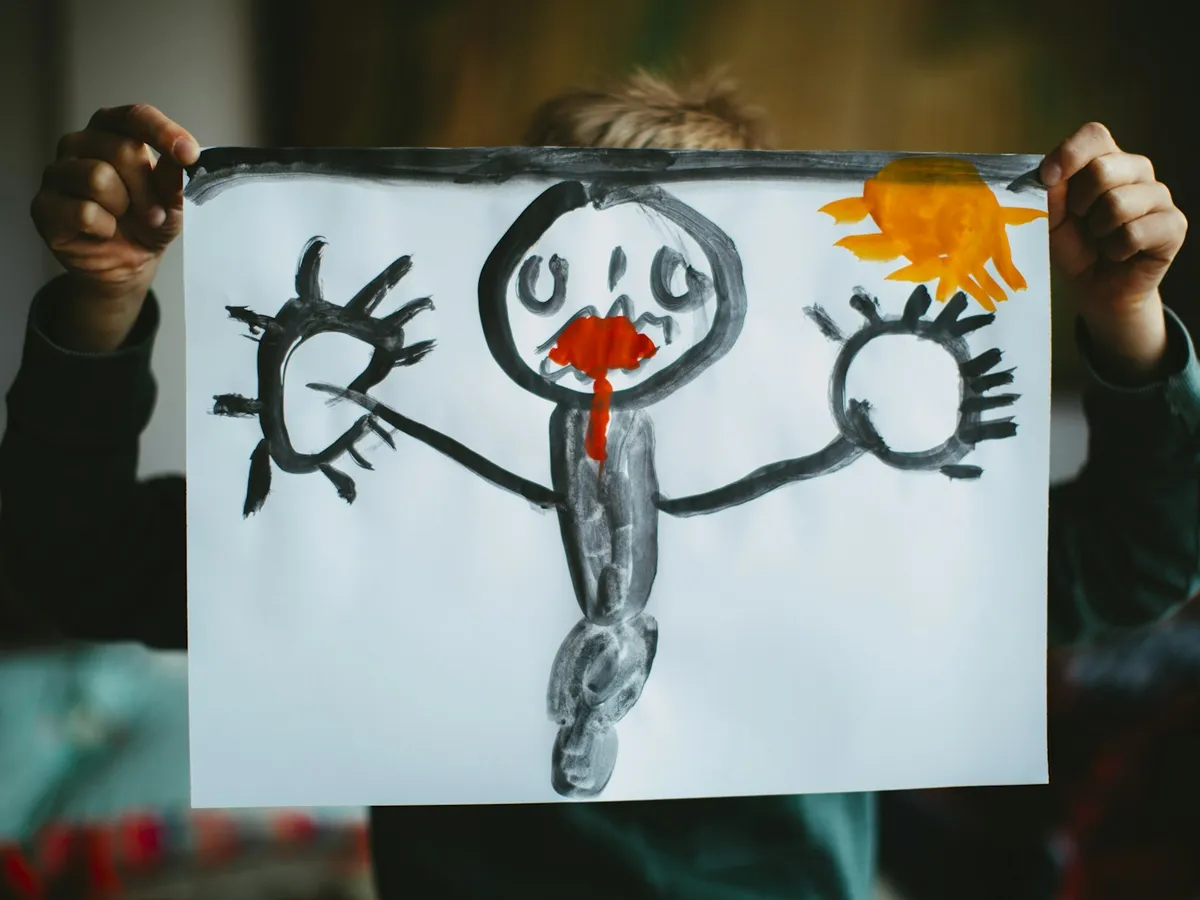 A toddler holding up a crayon drawing of a smiling person with big round hands