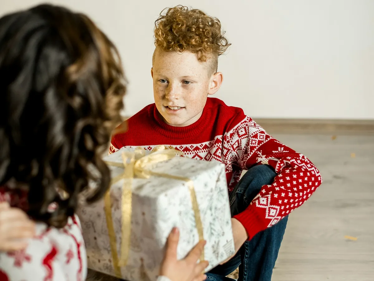 A boy in a Christmas sweater holding up a gold-wrapped present while a girl sits beside him